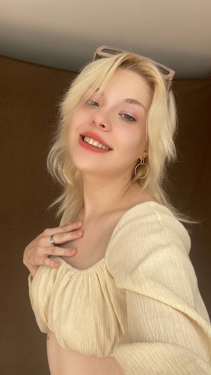 Sofia smiles in a cream off-the-shoulder top, sunglasses on head, hand on chest, against a brown background.