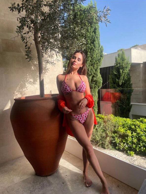 A barefoot woman in a pink polka-dot bikini and red shirt leans against a terracotta planter on a sunny stone patio.