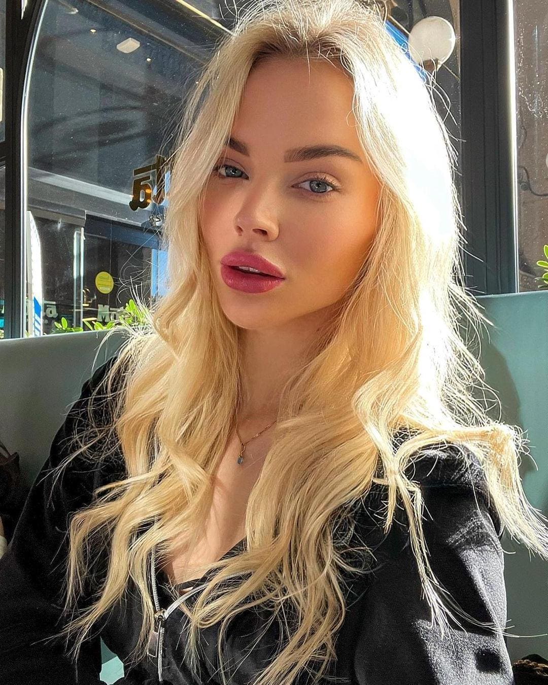 Seated in a booth, she wears a black zip-front top and thin necklace, with a window and greenery visible in the background.