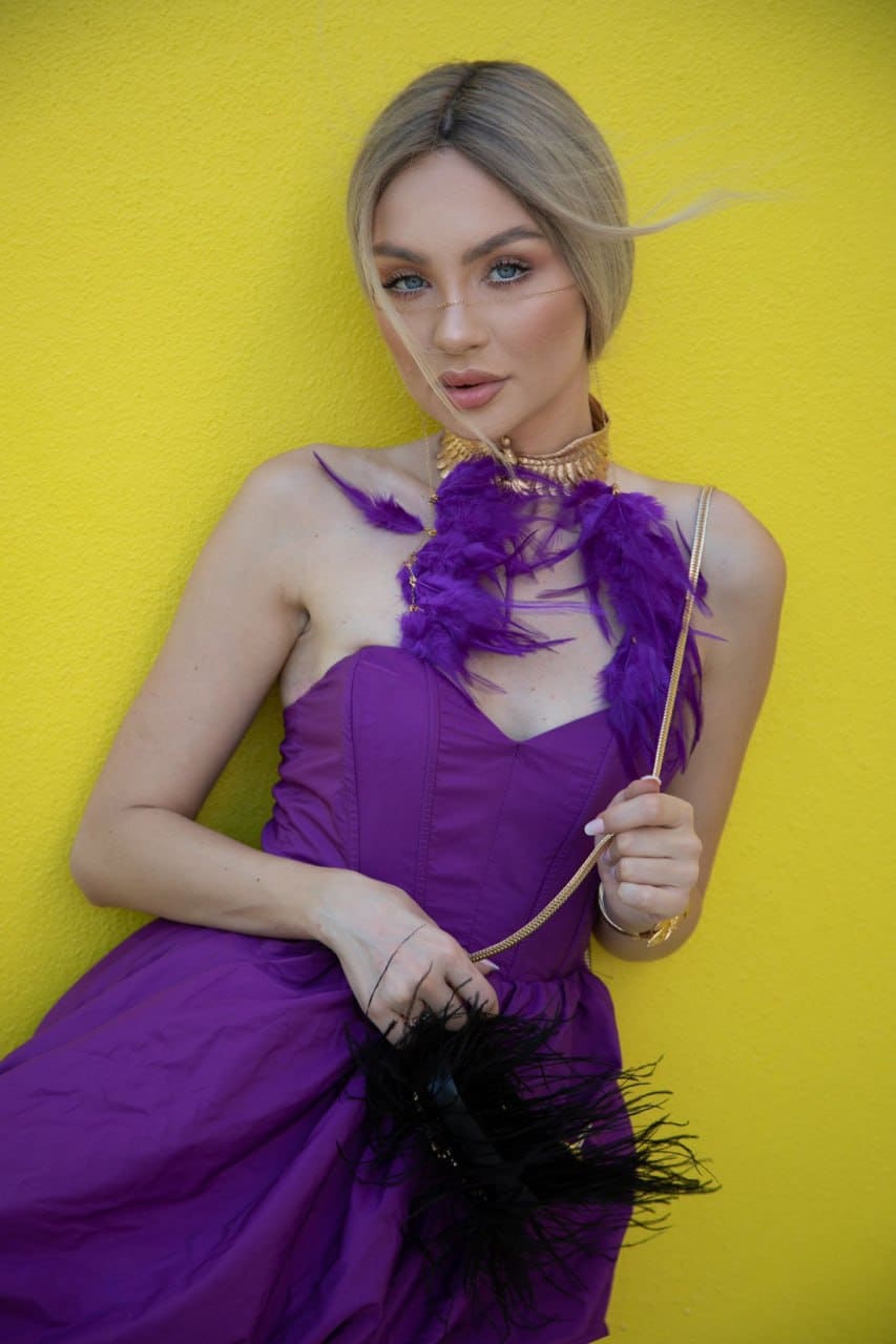 Bianca in a strapless purple dress with feathered neckline and gold choker, holding a black clutch against a yellow wall.