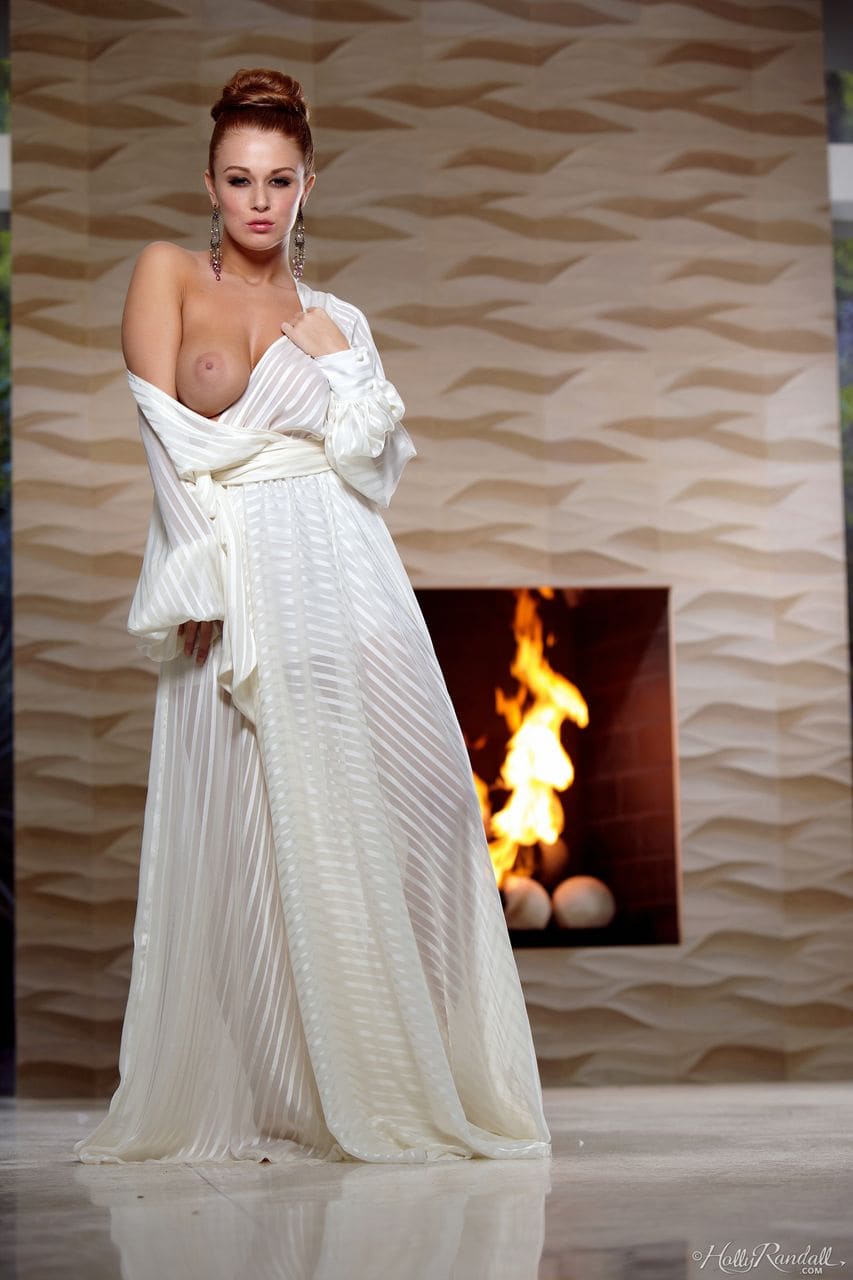 A woman in a sheer white robe stands before a lit fireplace, the garment pulled down to expose one breast.