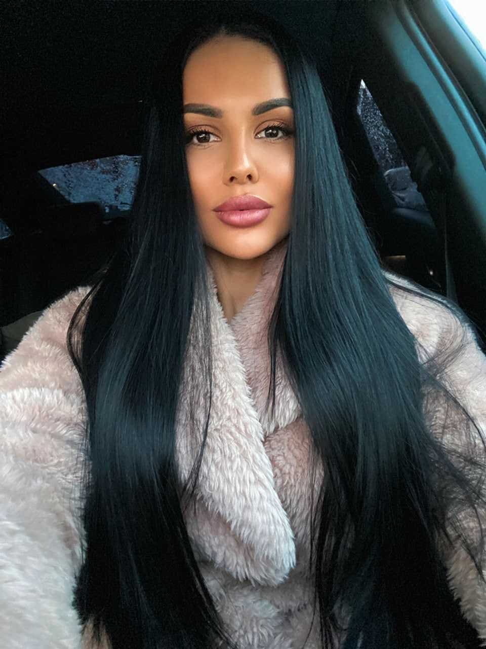 A close-up portrait of a woman inside a car, wearing a light-colored fluffy faux-fur coat with a wide collar.
