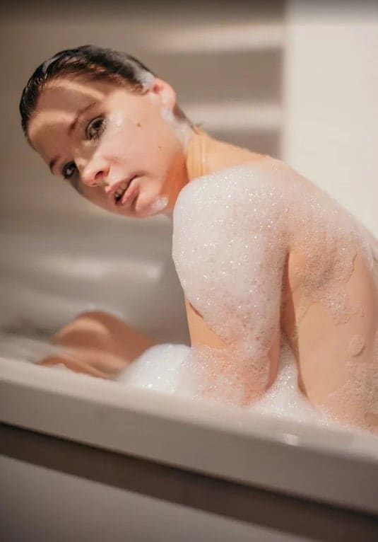 A woman with wet dark hair sits in a white bathtub filled with bubbles, her shoulders covered in foam as she looks toward the camera.