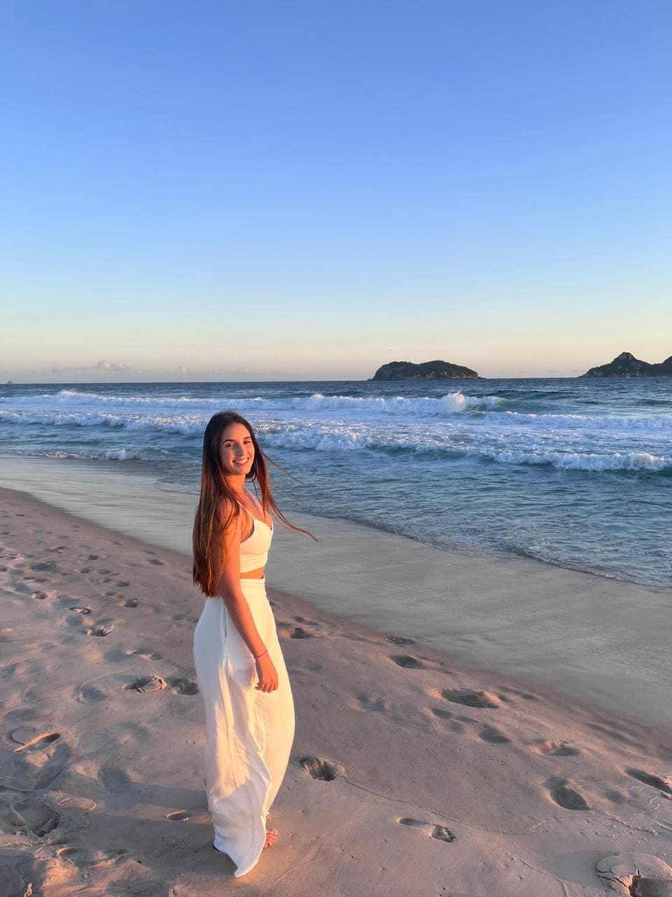 A woman stands barefoot on a beach in a white cropped top and long skirt, with ocean waves and islands behind.
