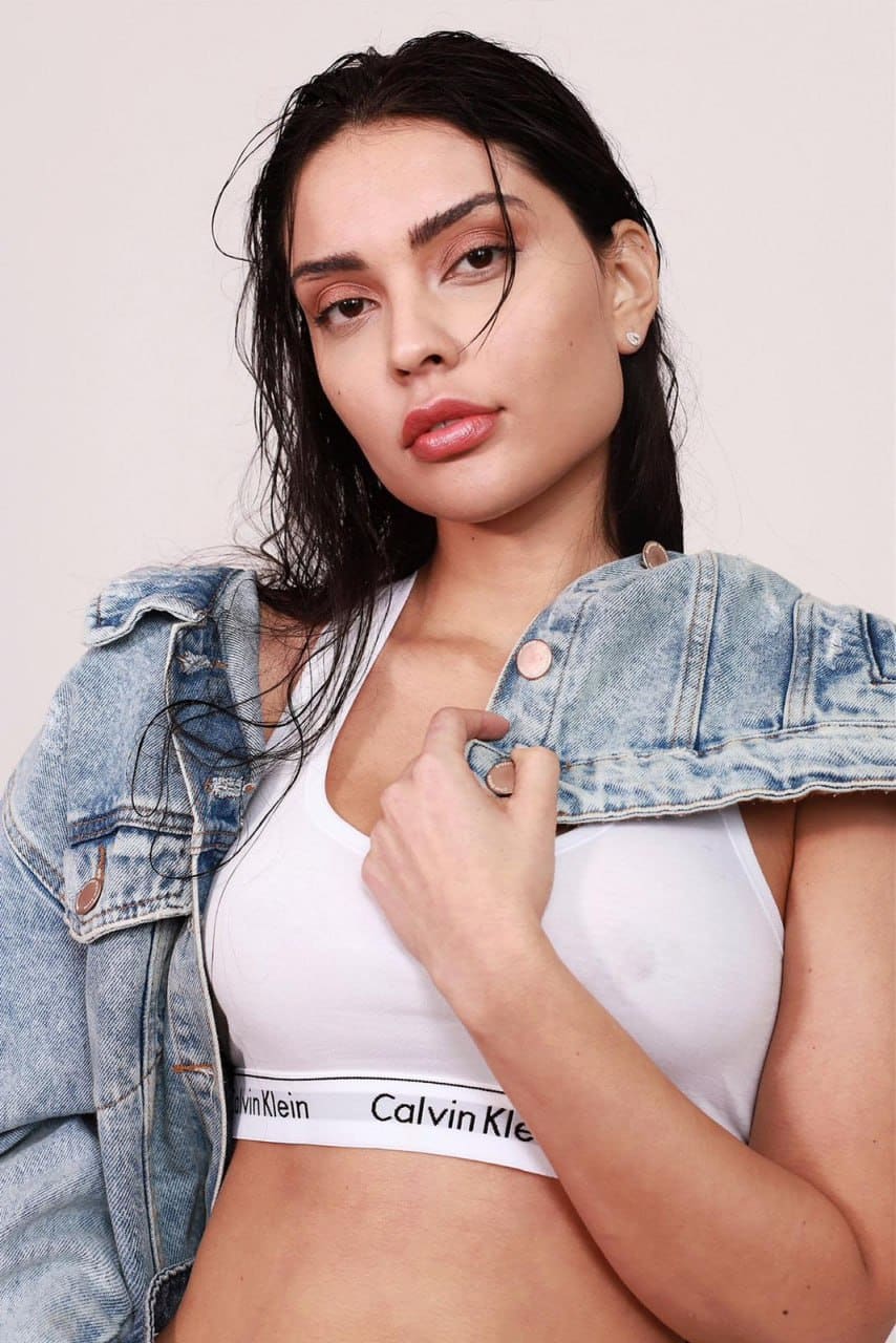 Against a plain light background, a woman with wet hair wears a white sports bra and holds an unbuttoned denim jacket draped over her shoulders.