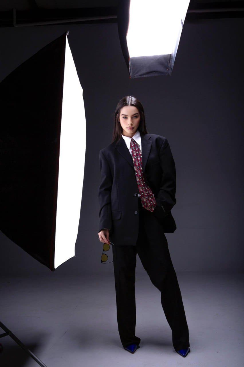 A woman stands in a studio in a black suit, white shirt, and red patterned tie, holding sunglasses while blue-tipped shoes are visible.