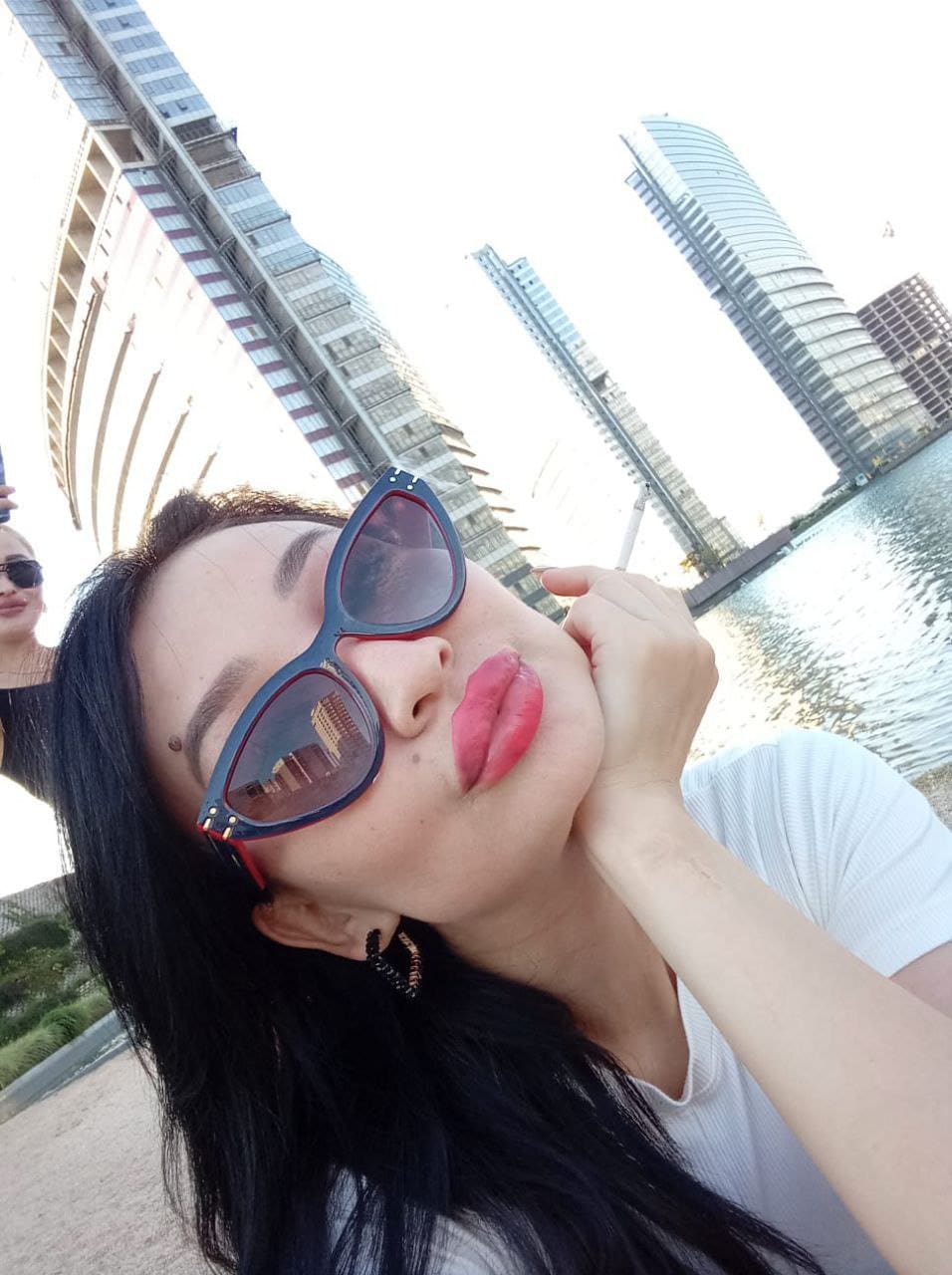 A woman in sunglasses and a white top poses near waterfront buildings, hand resting near her chin.