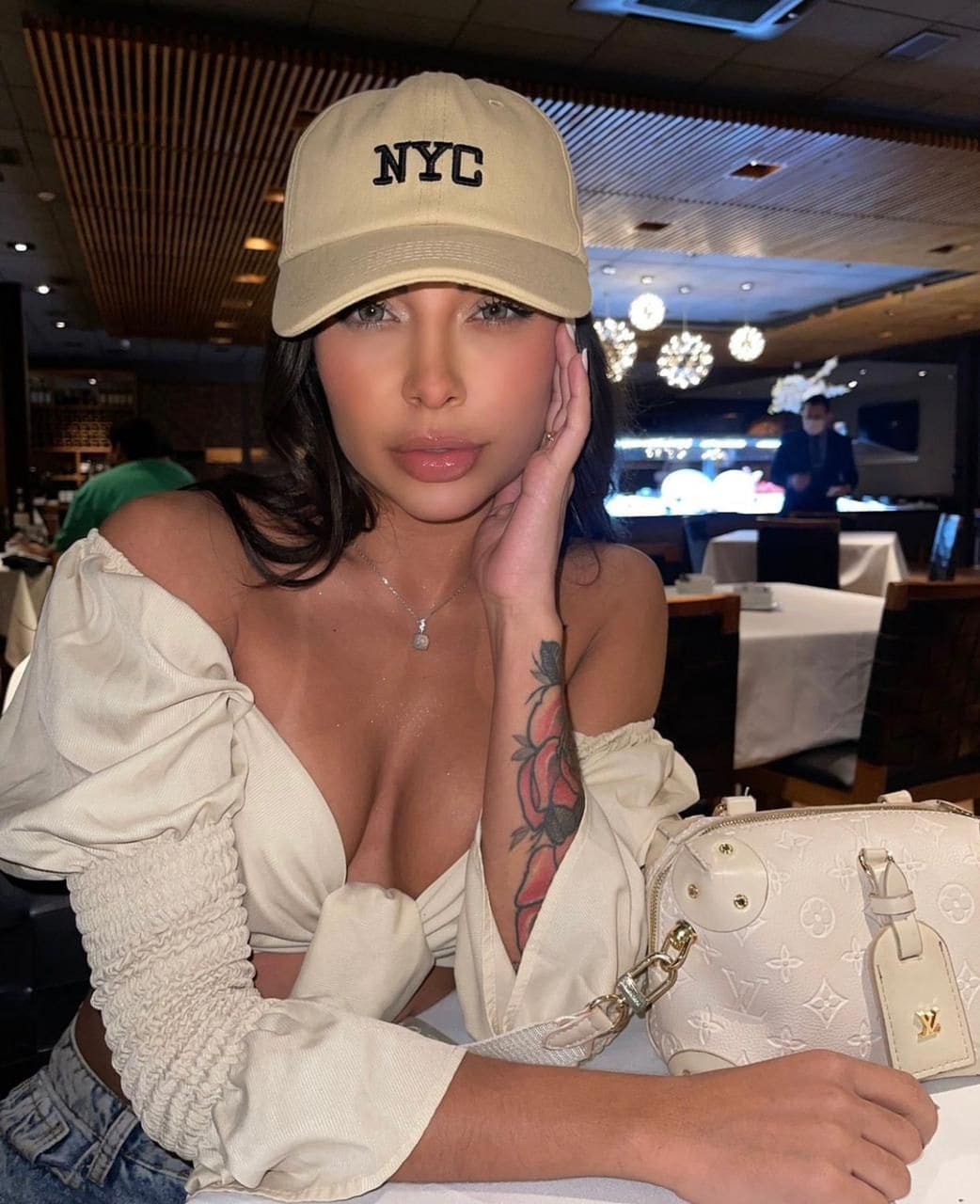 A woman in a beige cap and off-the-shoulder cream blouse rests her head on her hand at a restaurant table with a white bag.