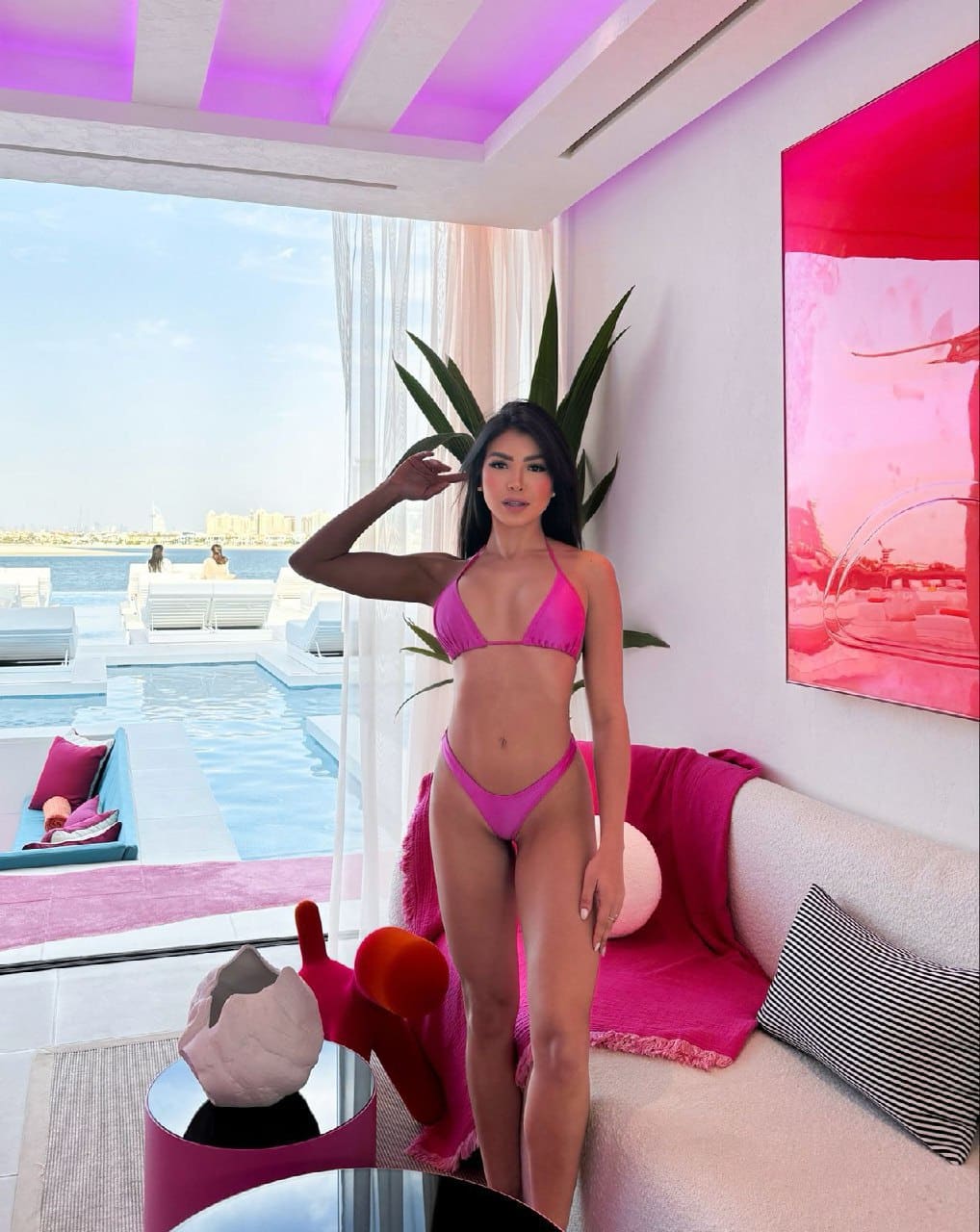 A woman in a pink bikini stands indoors near a pool, posing with one hand raised near her head in bright light.