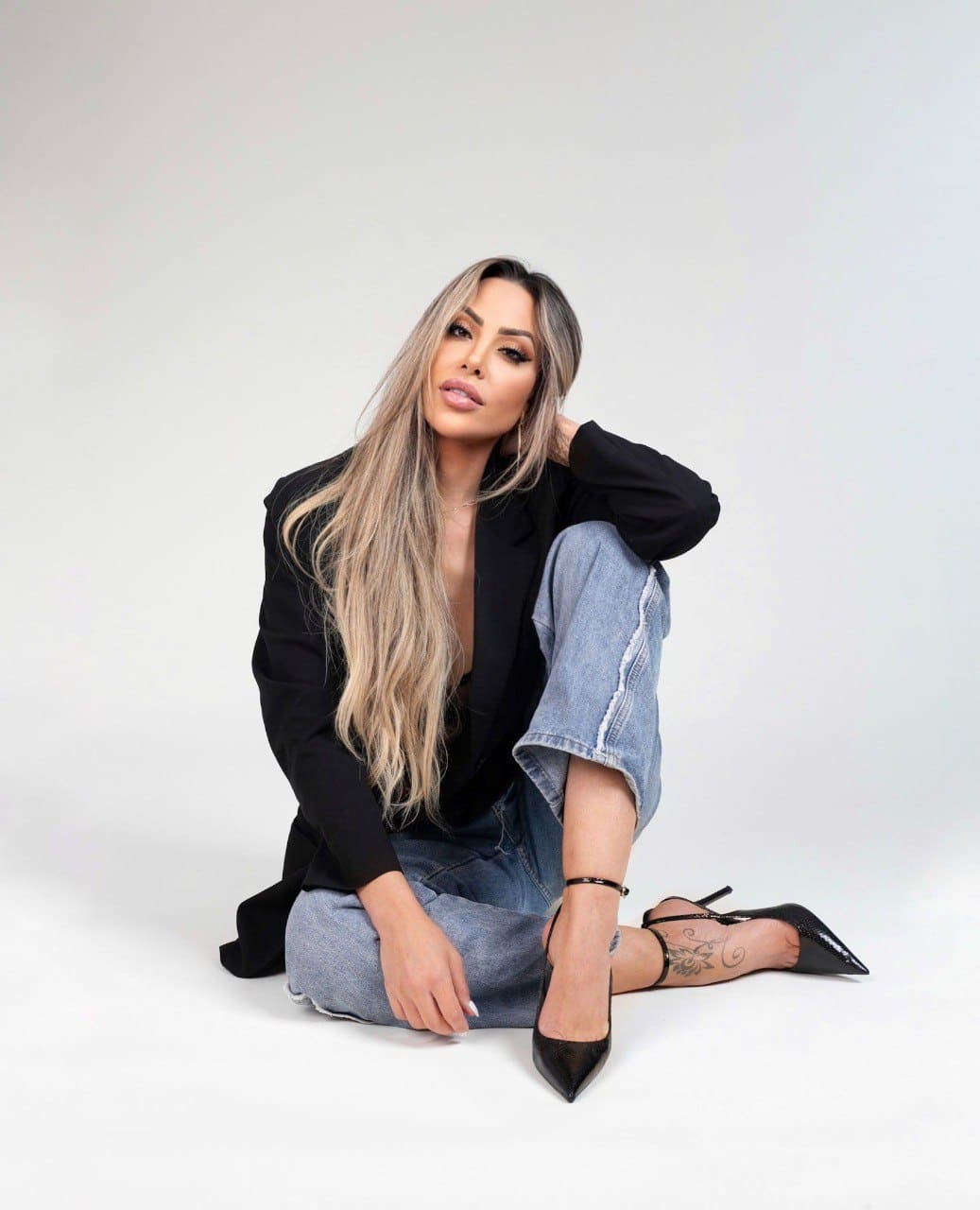 Dana sits on a white studio floor in a black blazer, light jeans, and heels, with one hand behind her head.