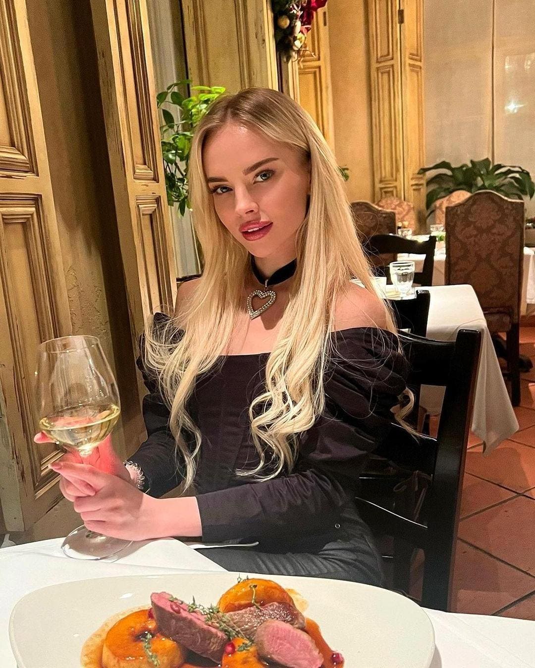 Seated at a restaurant table, she holds white wine in a black off-the-shoulder top and heart choker, with food plated ahead.