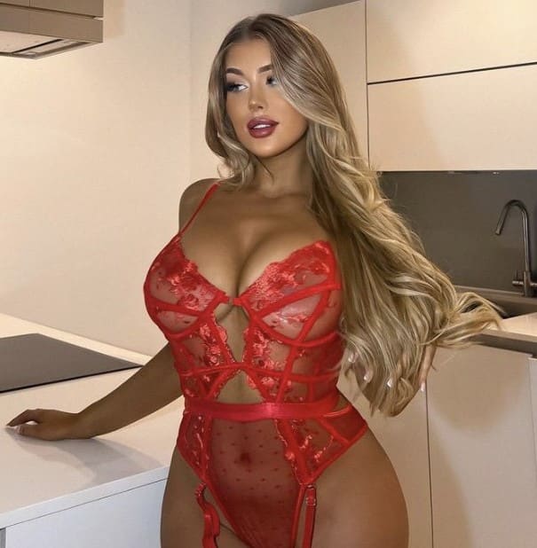 A blonde woman with wavy hair stands in a modern kitchen wearing a red lace and mesh bodysuit with cutouts, one hand on a white countertop.