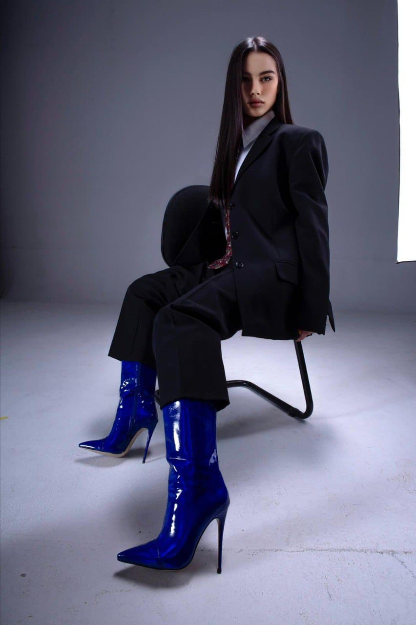 A woman sits on a black studio chair wearing a black suit jacket and trousers over a white top, with glossy blue high-heeled boots.
