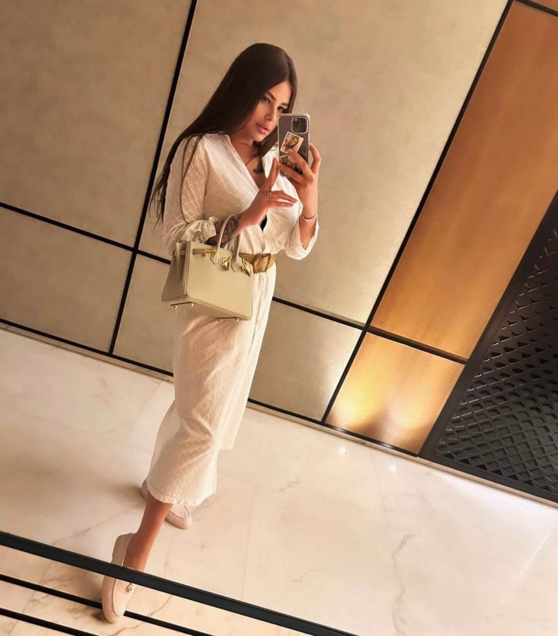 Standing before a mirror in a white textured dress with a gold belt, she holds a structured handbag in a room with marble floors.