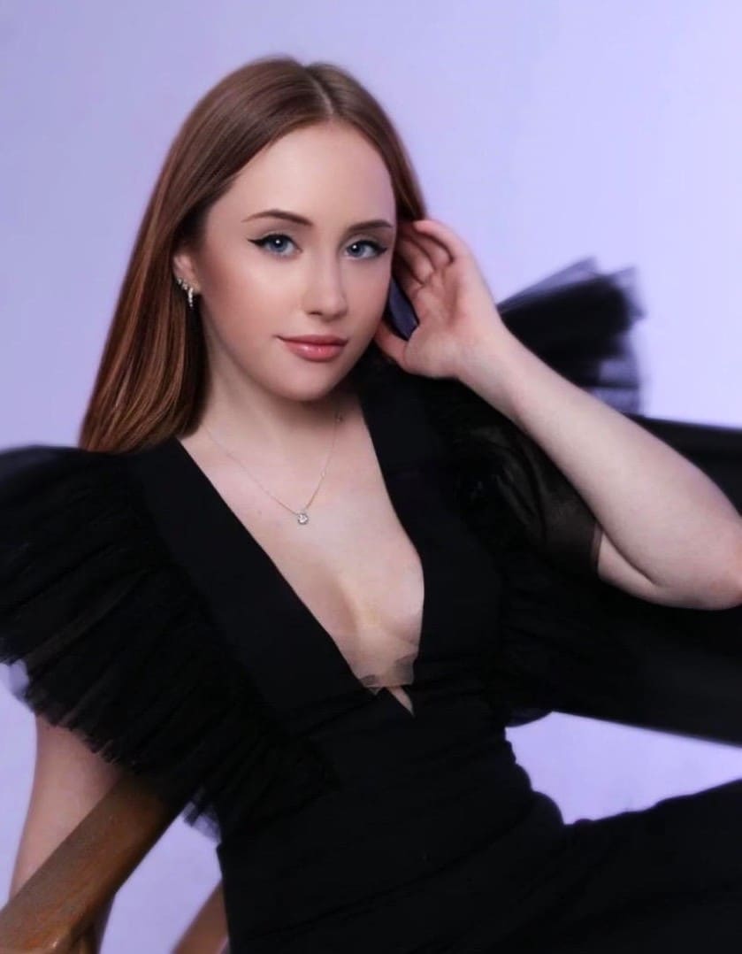 A seated woman in a black dress with sheer sleeves touches her hair while sitting in a wooden chair.