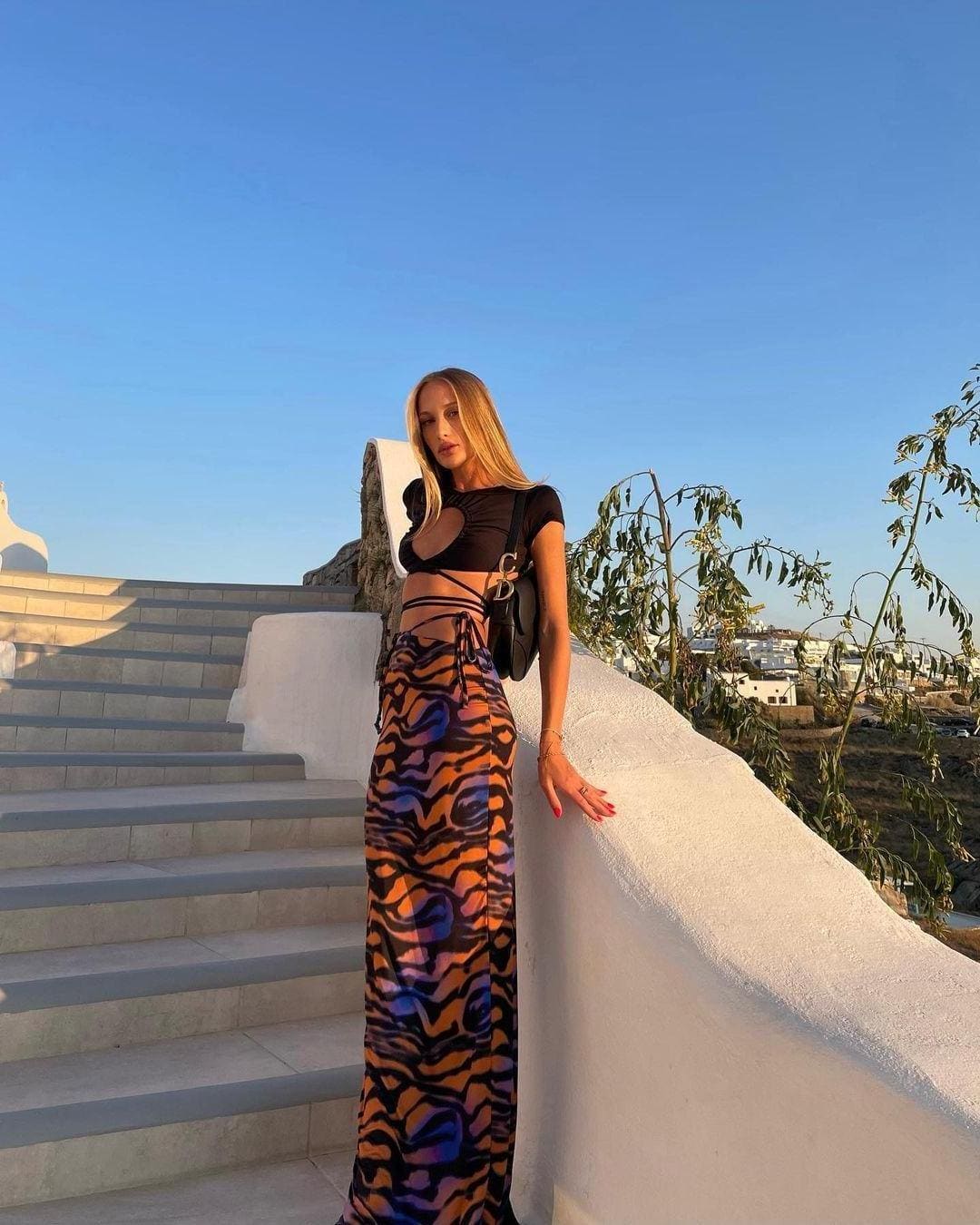 A woman stands on a white stone terrace near a staircase, wearing a black cut-out crop top, a long orange and purple skirt, and a small black bag.