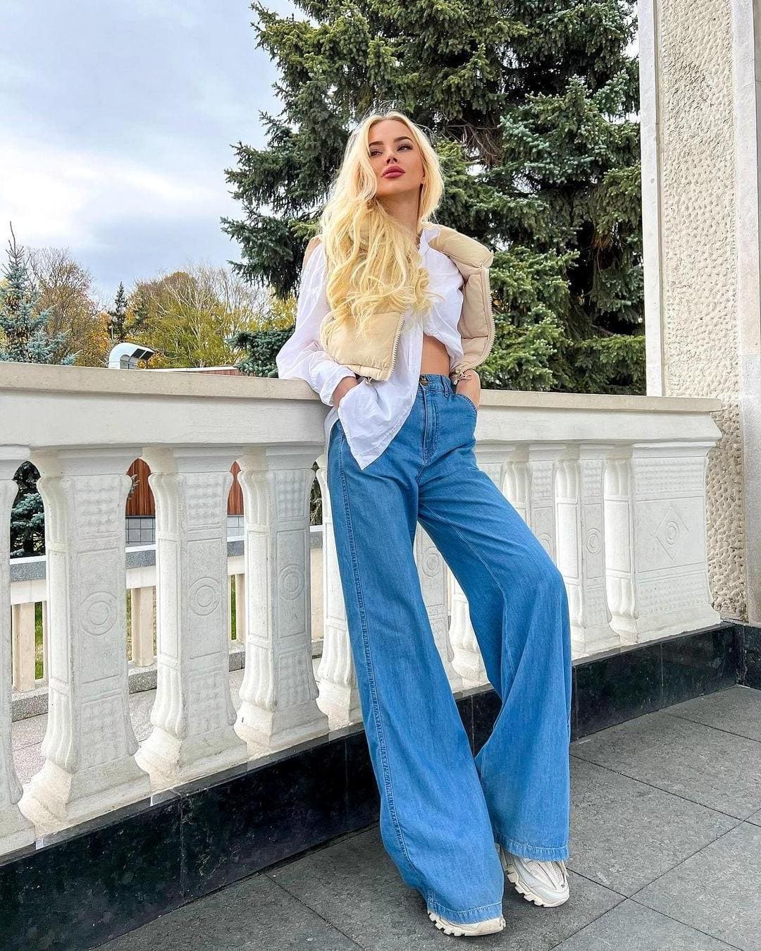 Leaning on a white balustrade outdoors, she wears a white crop, beige vest, blue jeans, and sneakers, with trees behind.