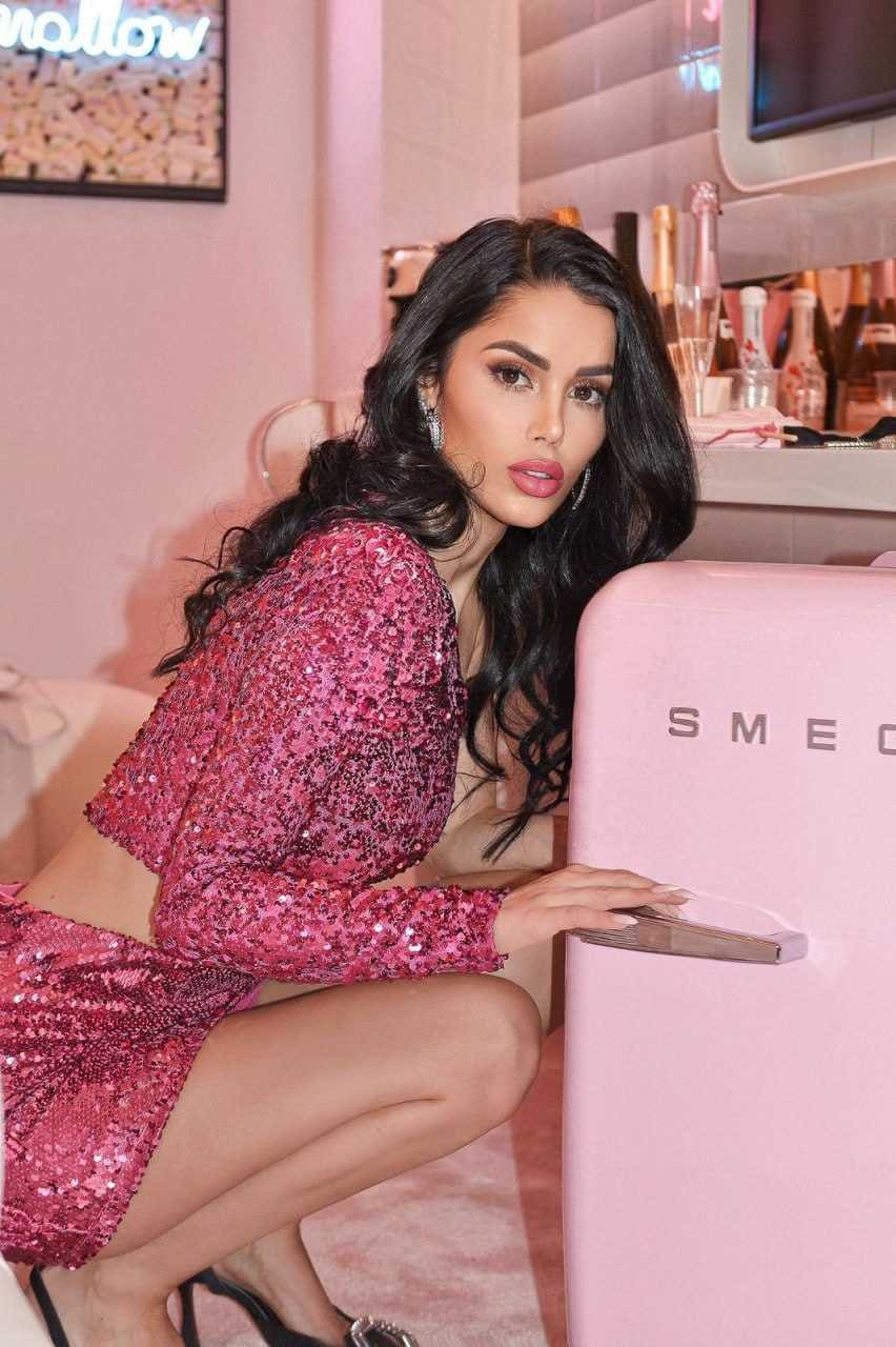 A woman in a pink sequined crop top and skirt crouches beside a pink retro refrigerator, hand on the handle, in a pink-walled room.