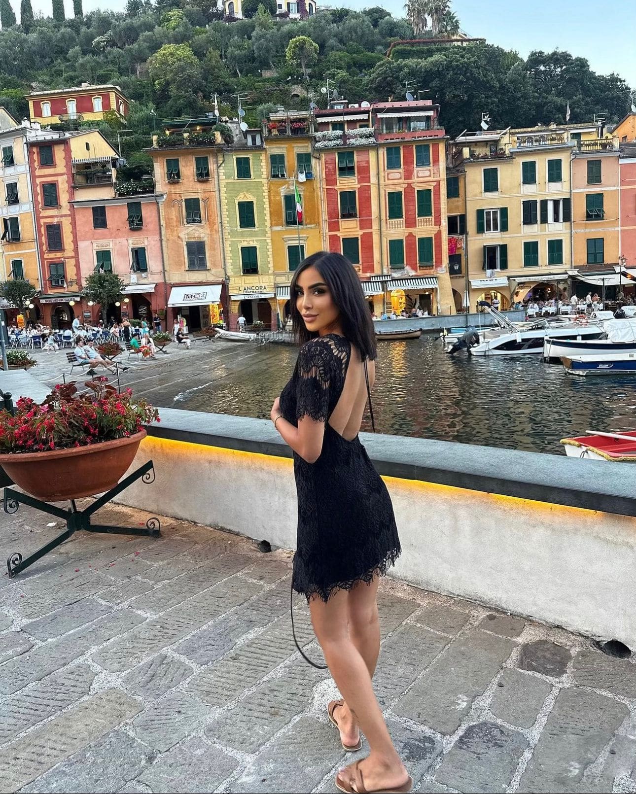 On a waterfront promenade, she wears a short black lace dress with an open back, with harbor buildings behind.