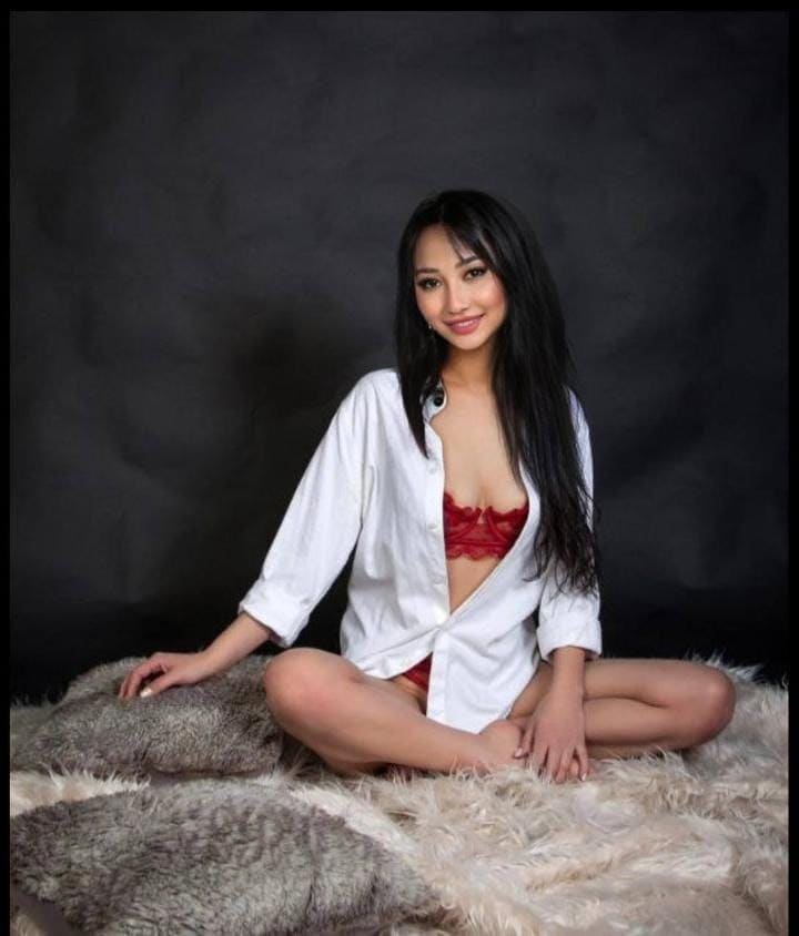 Nora sits cross-legged on a fur rug, wearing an open white shirt over red lace lingerie against a dark backdrop.