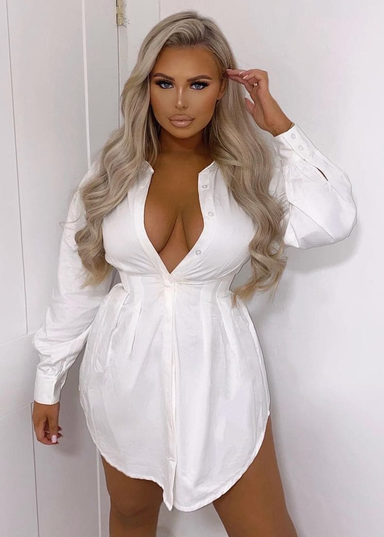 Paulina stands against a white paneled wall in a short white button-up shirt dress, raising one hand to her wavy blonde hair.