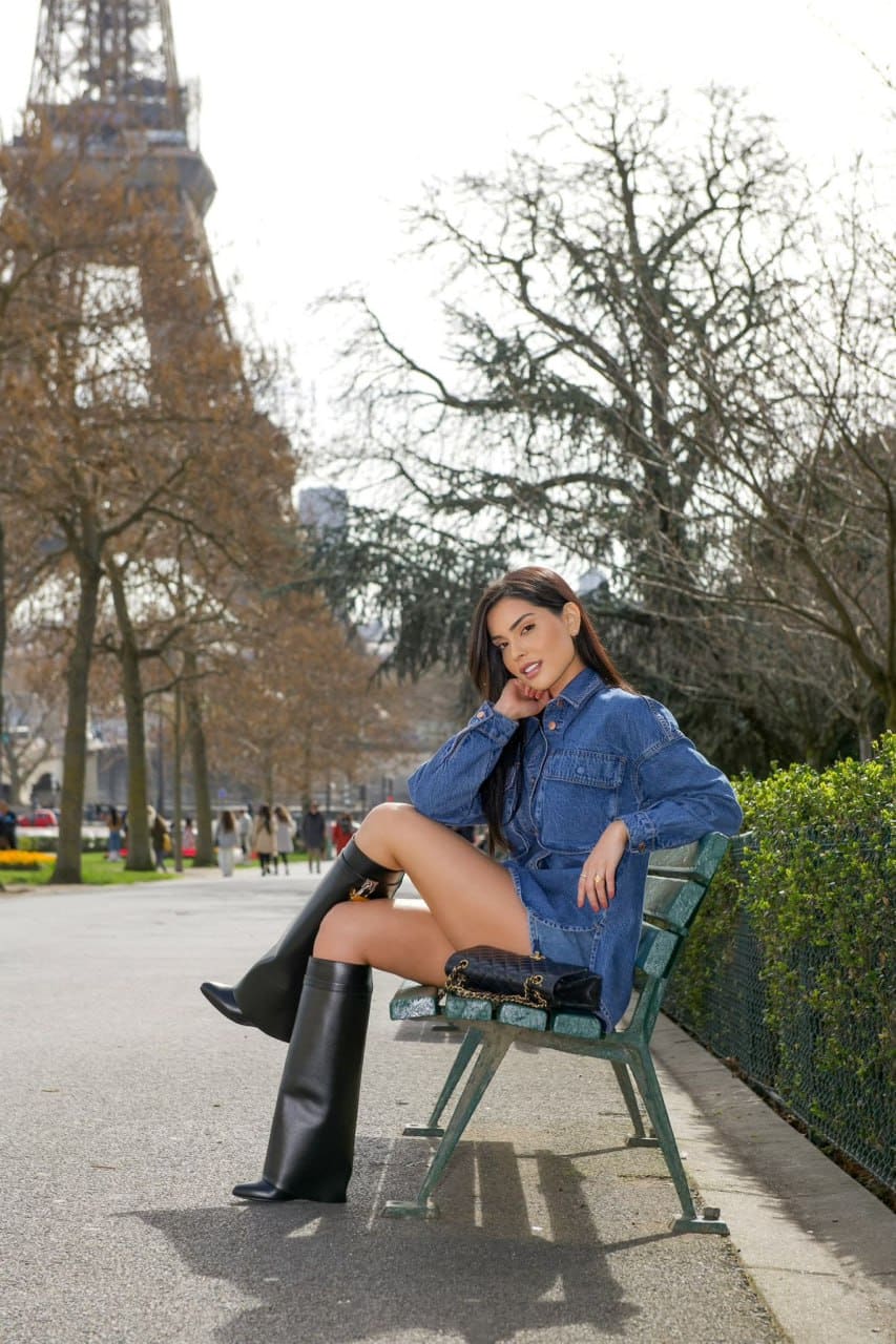 Seated on a green park bench, a woman in a denim shirt dress and black boots crosses her legs, with a tower and bare trees in the background.