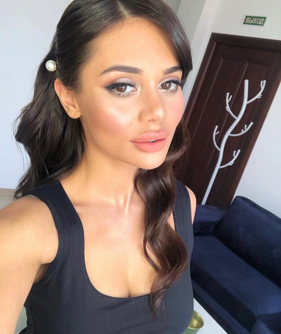 Her long wavy hair is held by a pearl clip as she wears a black sleeveless scoop-neck top, posing for a selfie near a dark wooden door and blue sofa.