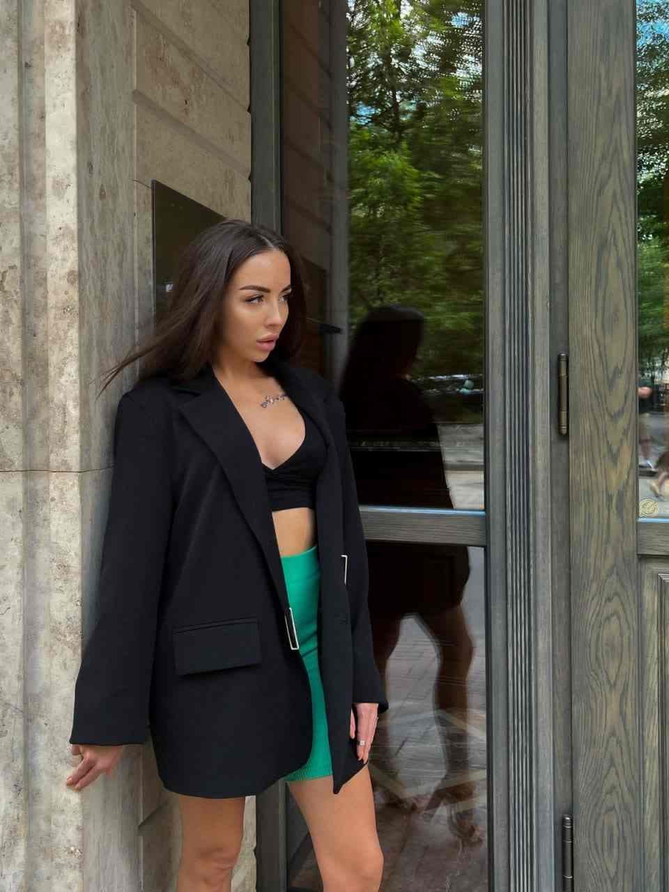 A woman leans against a stone pillar near a glass door in an open black blazer, black crop top, and green mini skirt, angled sideways.
