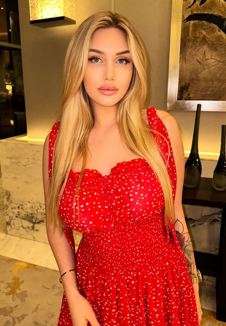 Amelia stands indoors in a red polka-dot dress with thin straps and a low neckline, showing a tattoo on her left arm.