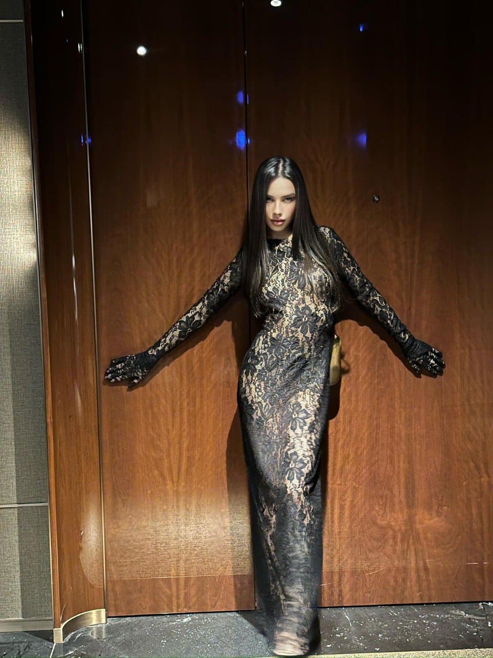 A woman stands with arms outstretched before a wooden wall, wearing a floor-length black lace dress with a high neckline and lace gloves.