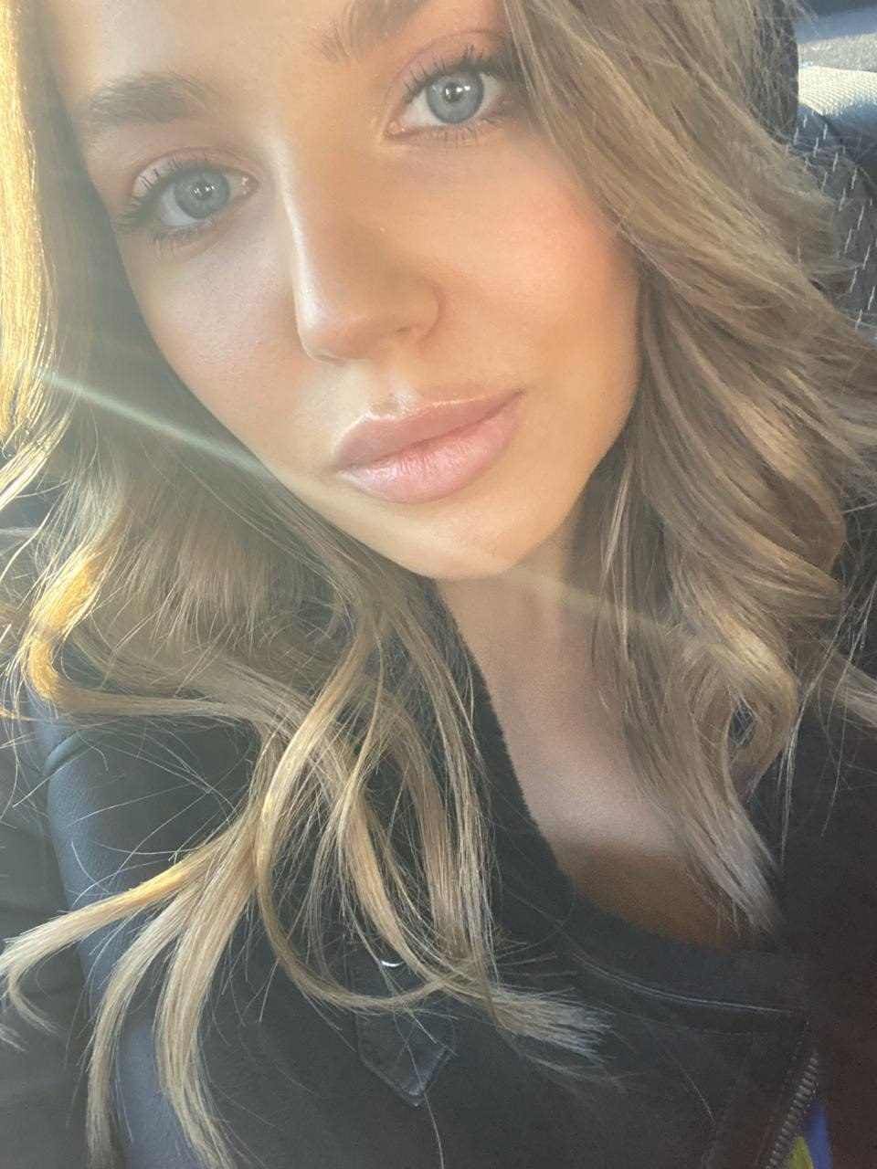 A blonde woman in a dark zippered jacket takes a selfie inside a car, with sunlight streaming in from the left.