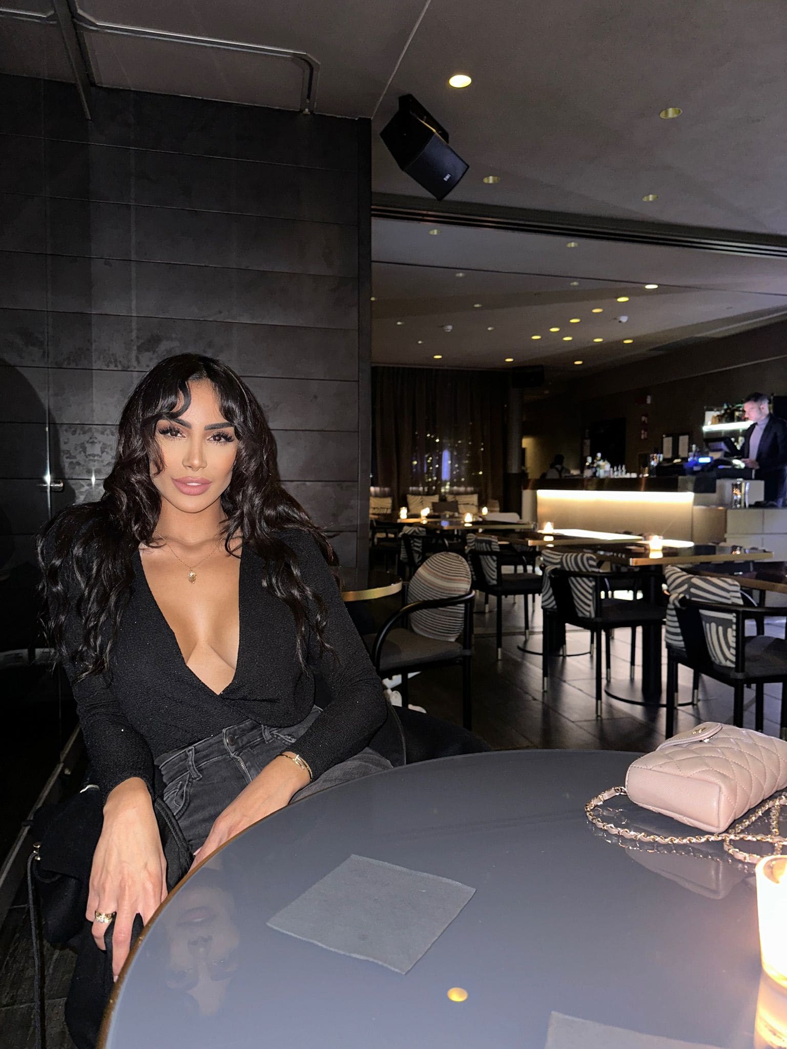At a round restaurant table, she wears a black V-neck top and dark jeans, with a small quilted bag in front.