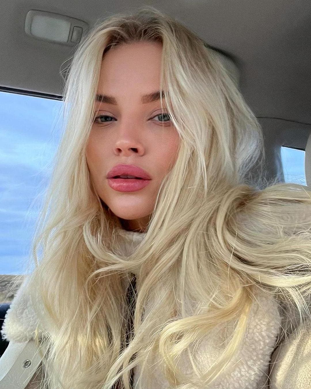 A close-up selfie shows her in a cream fleece jacket inside a car, with blue sky visible through the window behind her.