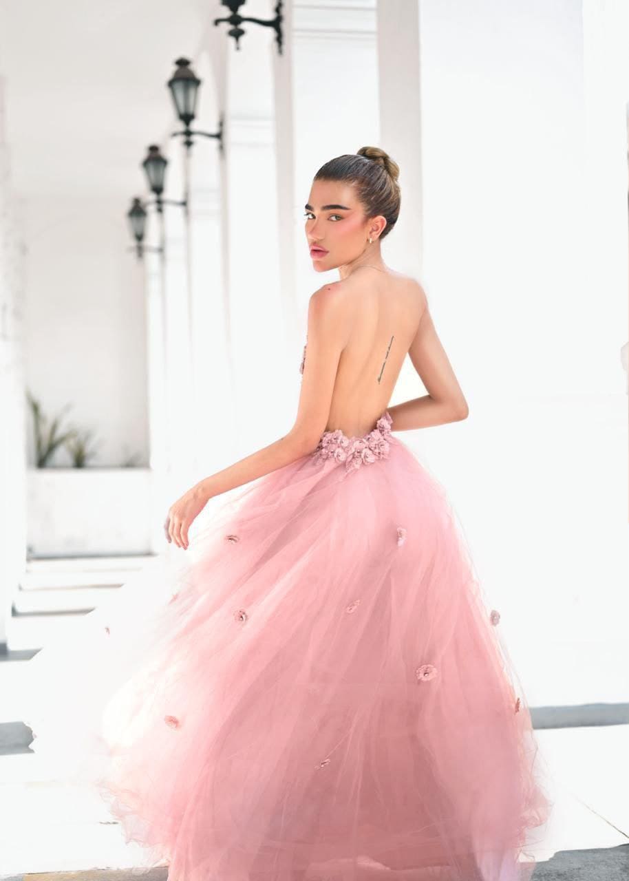 A dark-haired woman in a strapless pink tulle gown stands in a white corridor, looking over her shoulder toward the camera.
