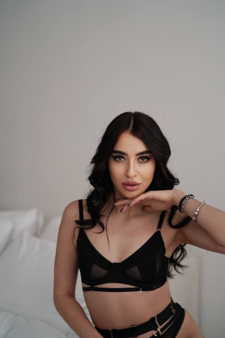 A woman in black strappy lingerie rests her chin on her hand, posing against a plain light wall near a white bed.