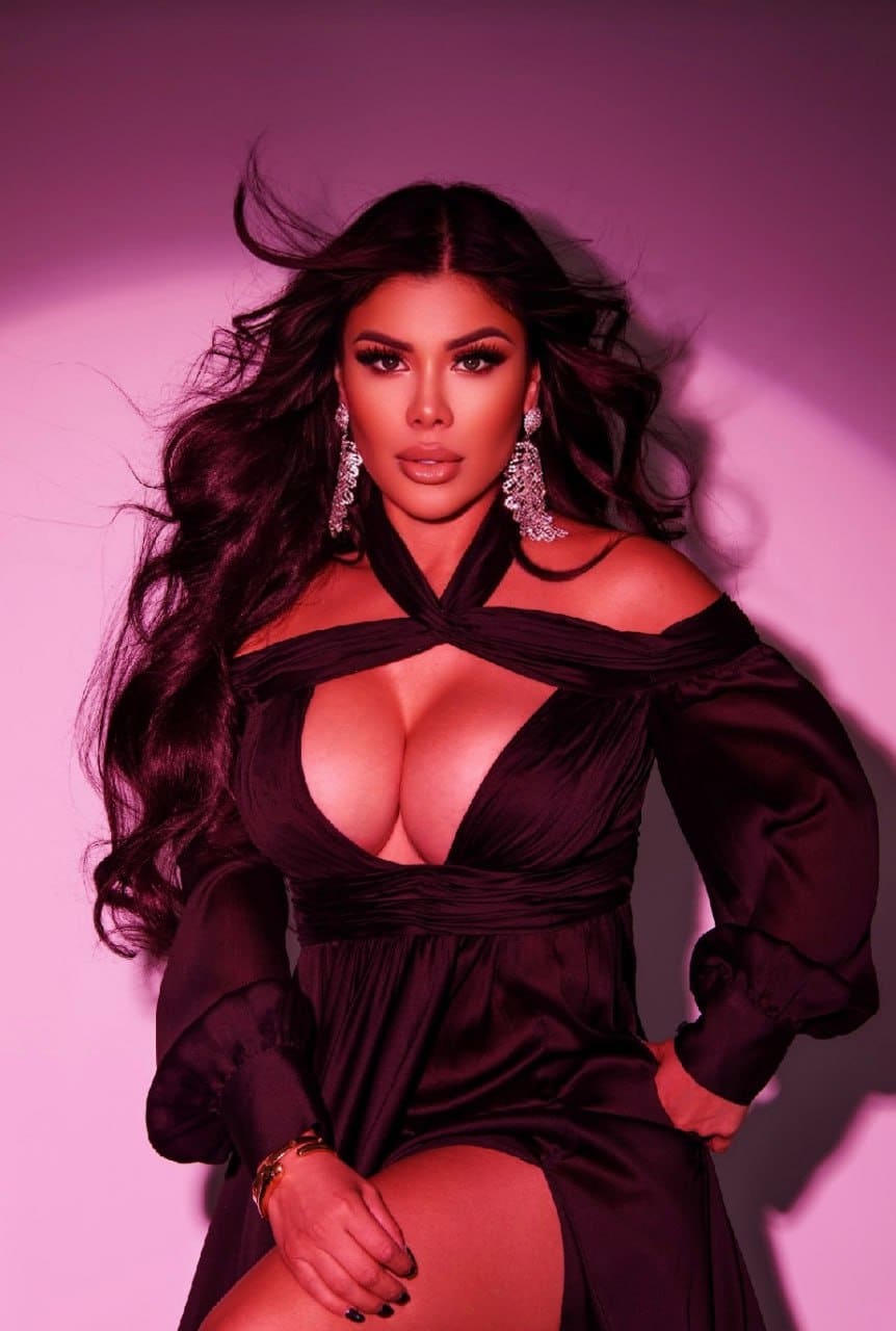 A woman in a black off-shoulder dress with a thigh slit stands against pink, wearing large earrings and a gold bracelet, hands on hip and thigh.