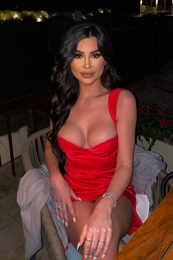 Vivienne sits in a woven chair outdoors at night, wearing a short red satin dress with a deep neckline and jewelry.