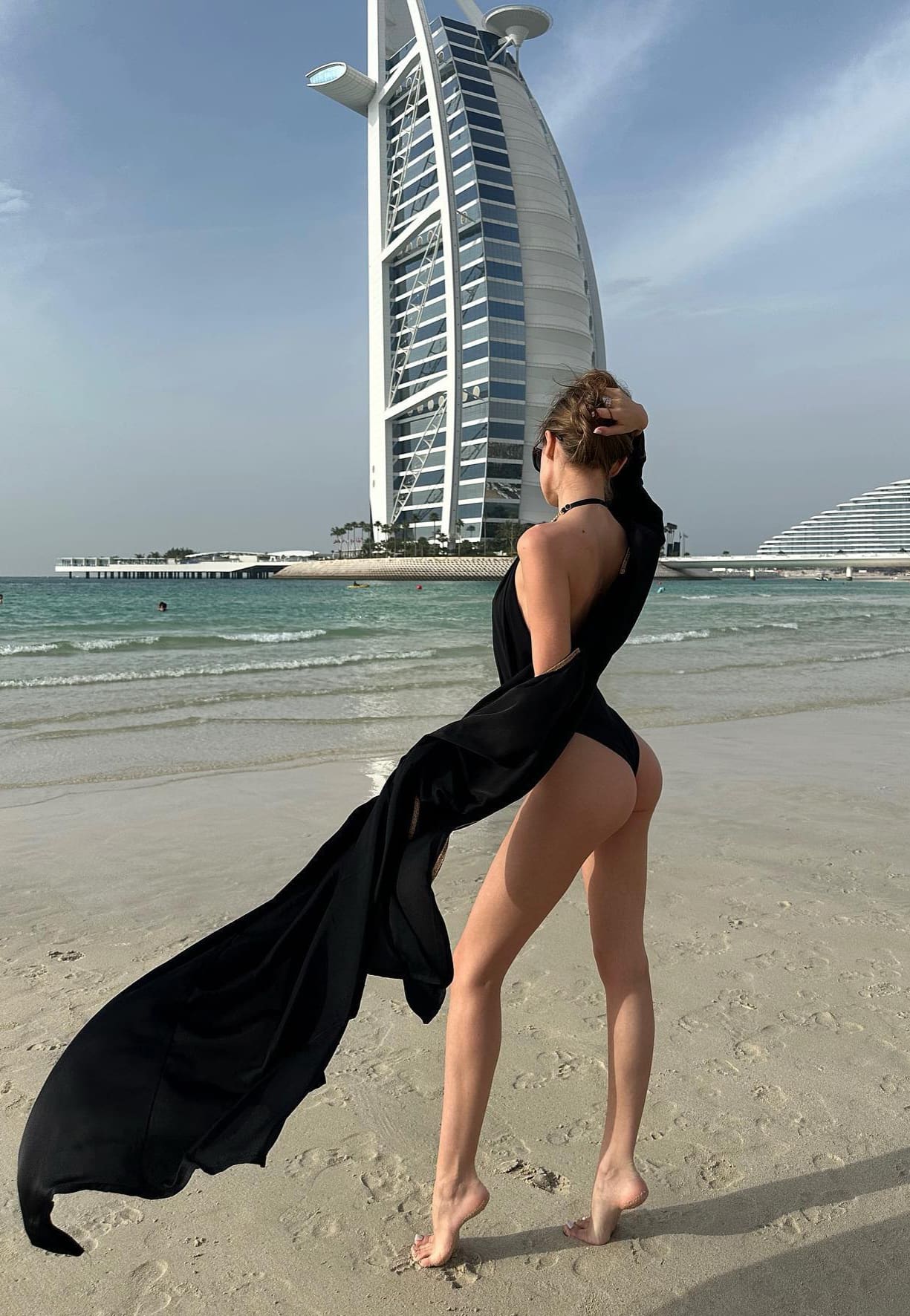 A woman in a black halter dress with a train stands barefoot on a beach, posing near water with a white sail-shaped hotel in the background.