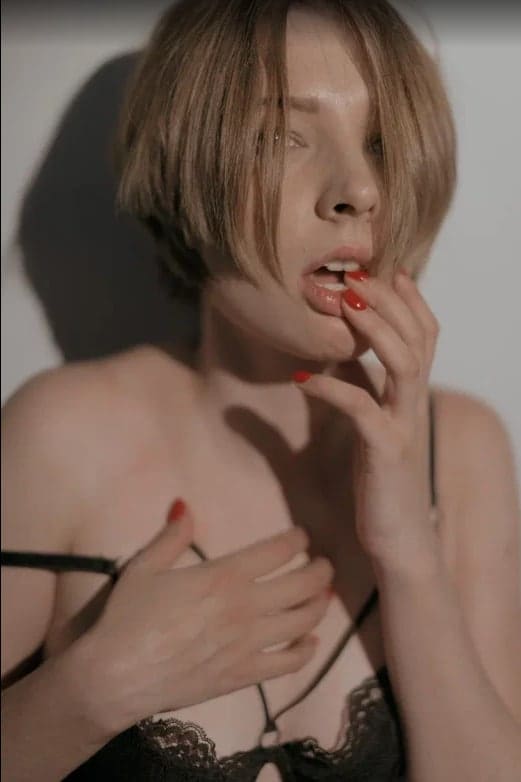 A person with short light-brown hair and red nails poses in a black lace bra, touching their mouth with one hand and resting the other on their chest.