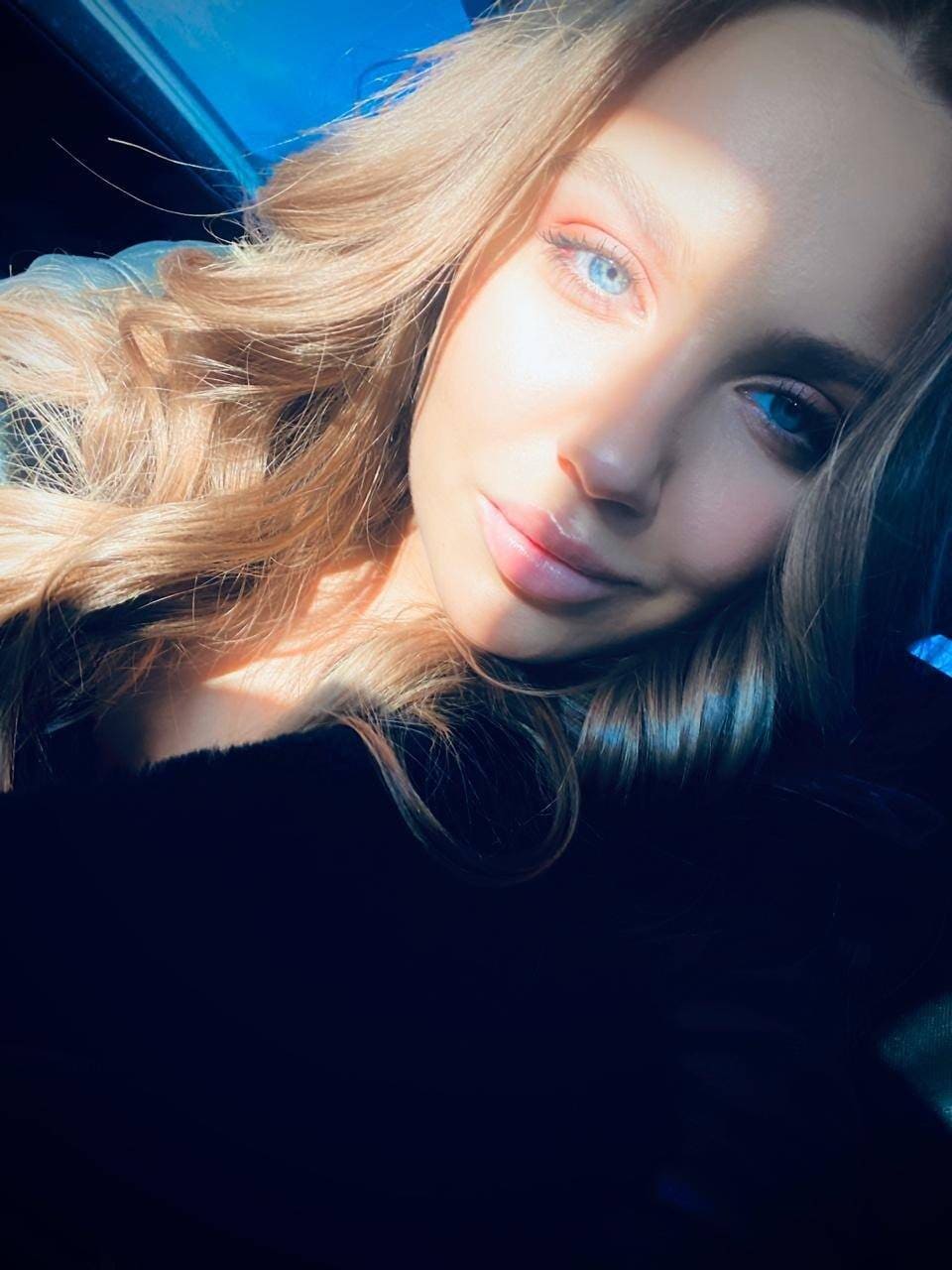 A close-up selfie shows a blonde woman in a dark top, with diagonal sunlight casting shadows across her face against a dark background.