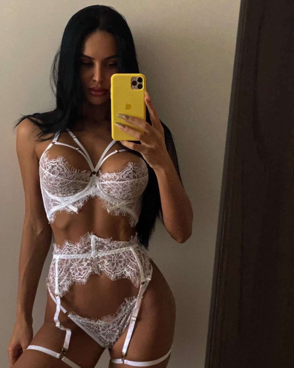 A woman takes a mirror selfie in a white lace lingerie set with garters, holding a yellow phone against a plain wall.