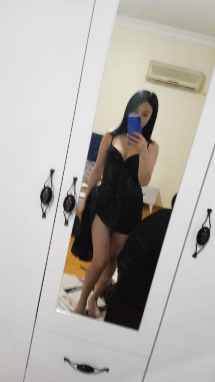 A barefoot woman in a black slip dress takes a mirror selfie with a blue phone in front of a wardrobe.