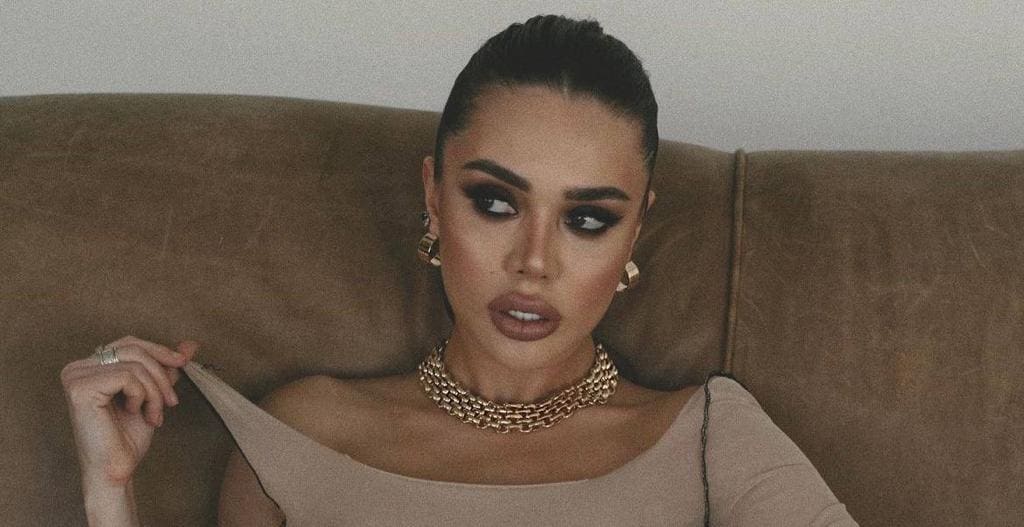 Sitting on a brown leather couch, she wears a beige off-shoulder top and chunky gold chain, pulling at the neckline with one hand while wearing gold earrings.