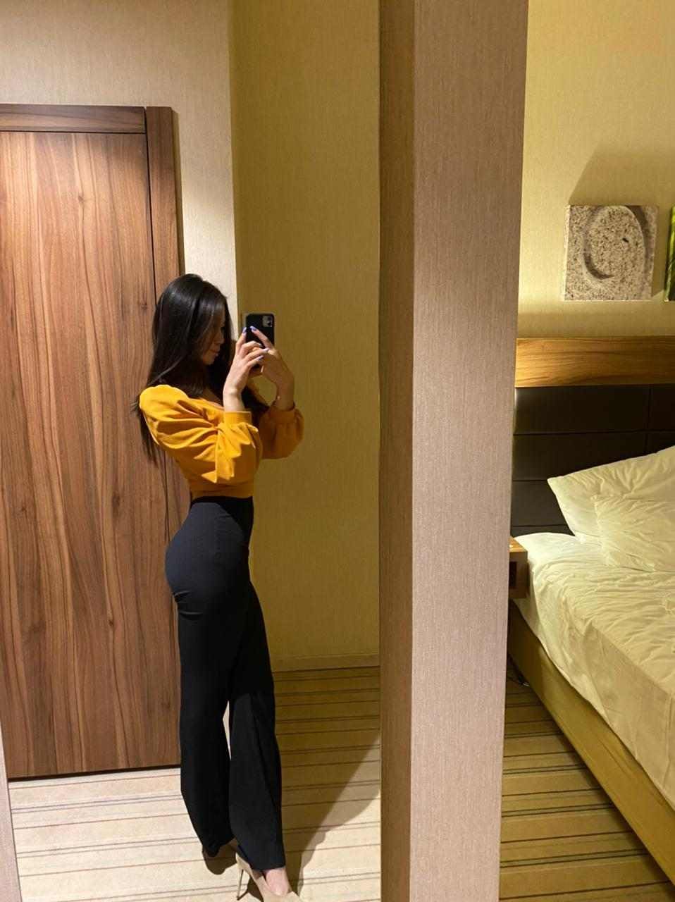 A mirror selfie shows a woman in a yellow long-sleeved top and black wide-leg pants, with a bed and wooden door visible in the bedroom background.