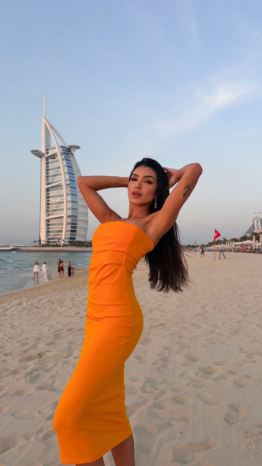 A woman stands on a beach in an orange strapless dress with hands behind her head, a sail-shaped skyscraper behind her.
