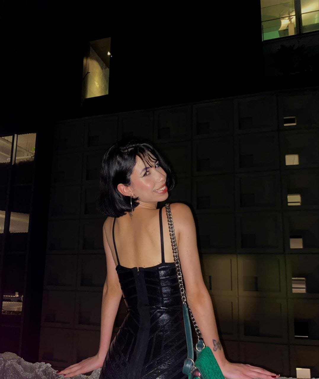 Facing away in a black strapless dress, she holds a green handbag against a dark building facade at night.