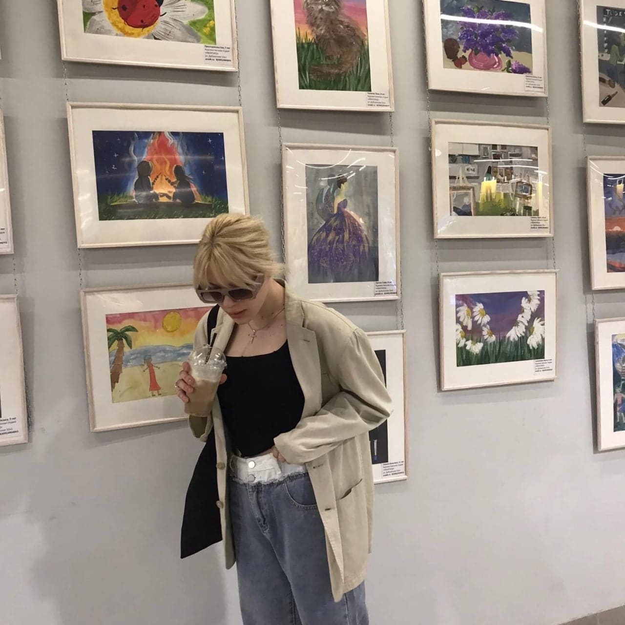 Person in beige blazer and jeans holds a drink in front of framed paintings, one hand in pocket.