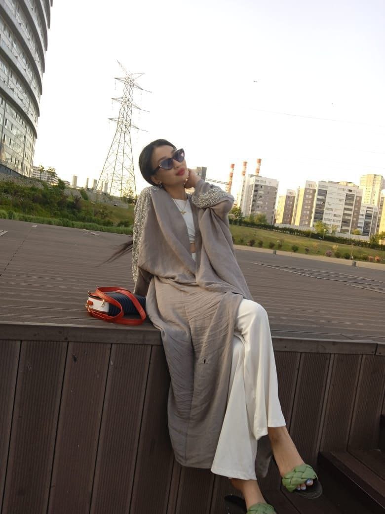 A seated woman in a grey cardigan and white pants wears sunglasses, with a handbag beside her in an urban setting.