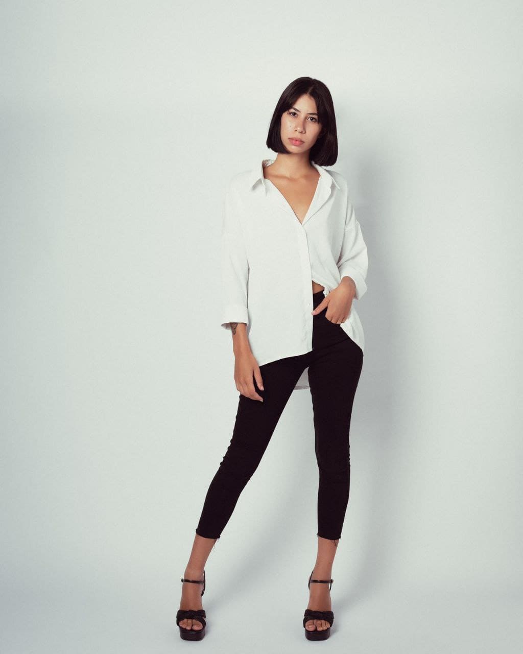 BIANCA stands against a light background in an unbuttoned white shirt, black cropped pants, and platform sandals, with one hand in her pocket.