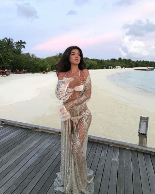 A woman in a sheer off-shoulder dress over a white bikini stands on a wooden deck, hands clasped near her chest.